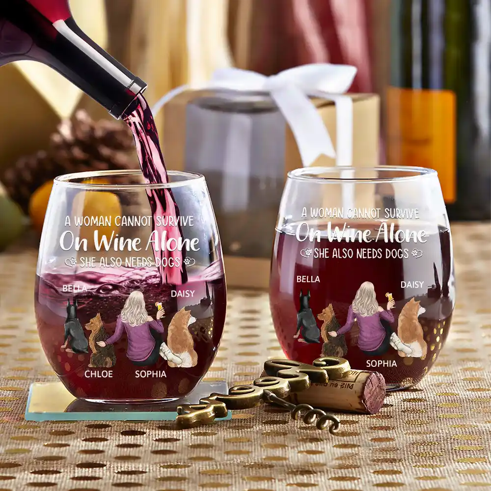 Two personalized wine glasses with red wine being poured into the left glass. The glasses feature an image of a woman surrounded by four dogs. The glasses are on a gold tablecloth with a decorative metal wine opener and cork. A gift box with a white ribbon is in the background.