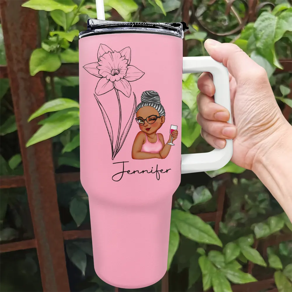 A hand holds a pink tumbler with a white handle, decorated with a cartoon woman, a daffodil, and the name "Jennifer." The tumbler has a lid with a straw. Green foliage is visible in the background.
