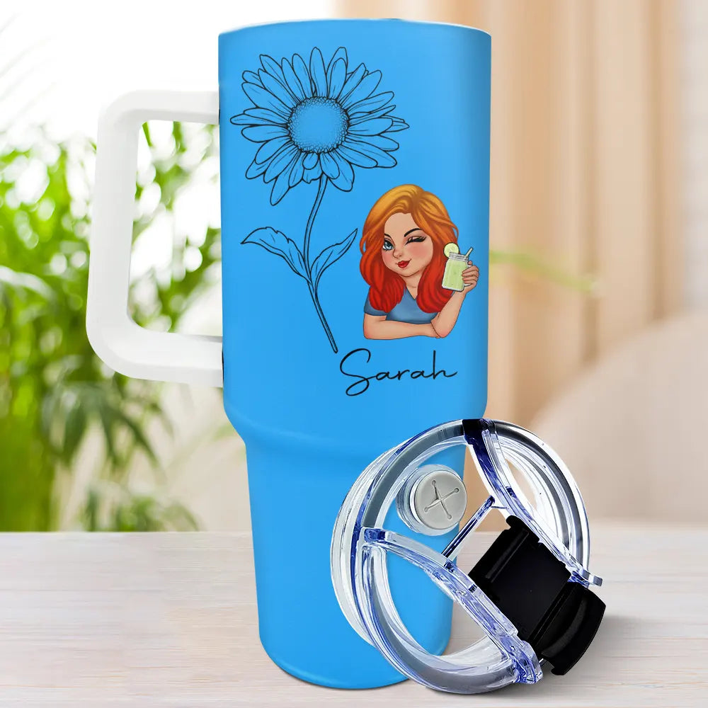 A light blue tumbler with a white handle, featuring a line art daisy, a cartoon woman with red hair holding a green drink, and the name "Sarah" in cursive, with a clear and black lid resting on the table next to it.