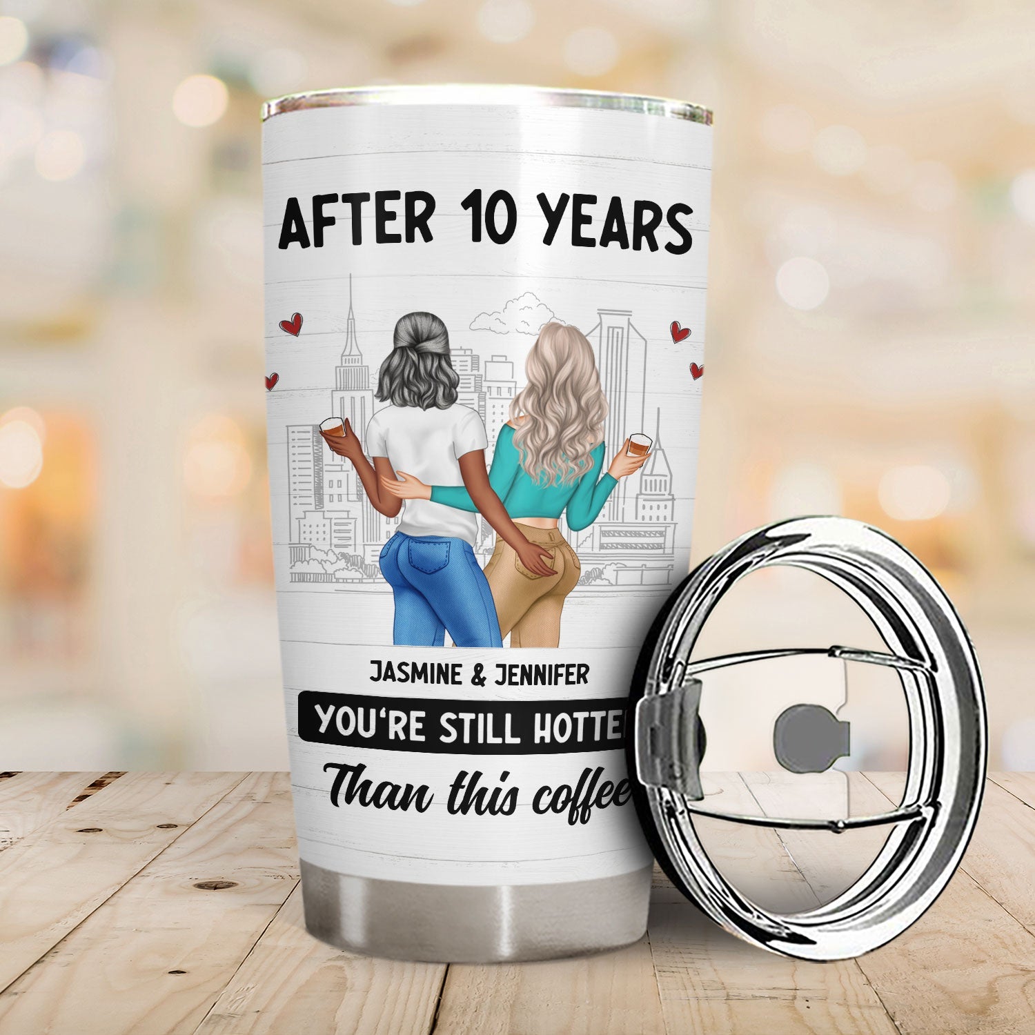 White stainless steel tumbler with cartoon illustration of two women, cityscape, and the words "After 10 years, Jasmine & Jennifer, You're Still Hotter Than this coffee", on a wooden surface with the lid beside it.