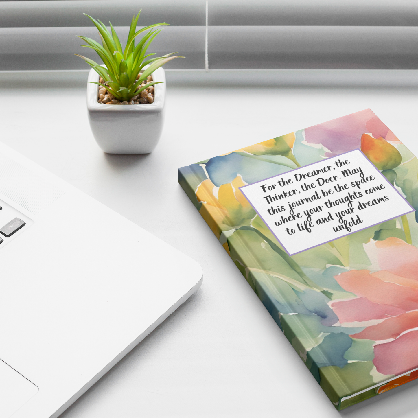 Floral watercolor journal with inspirational quote on cover beside white laptop and small green potted plant.