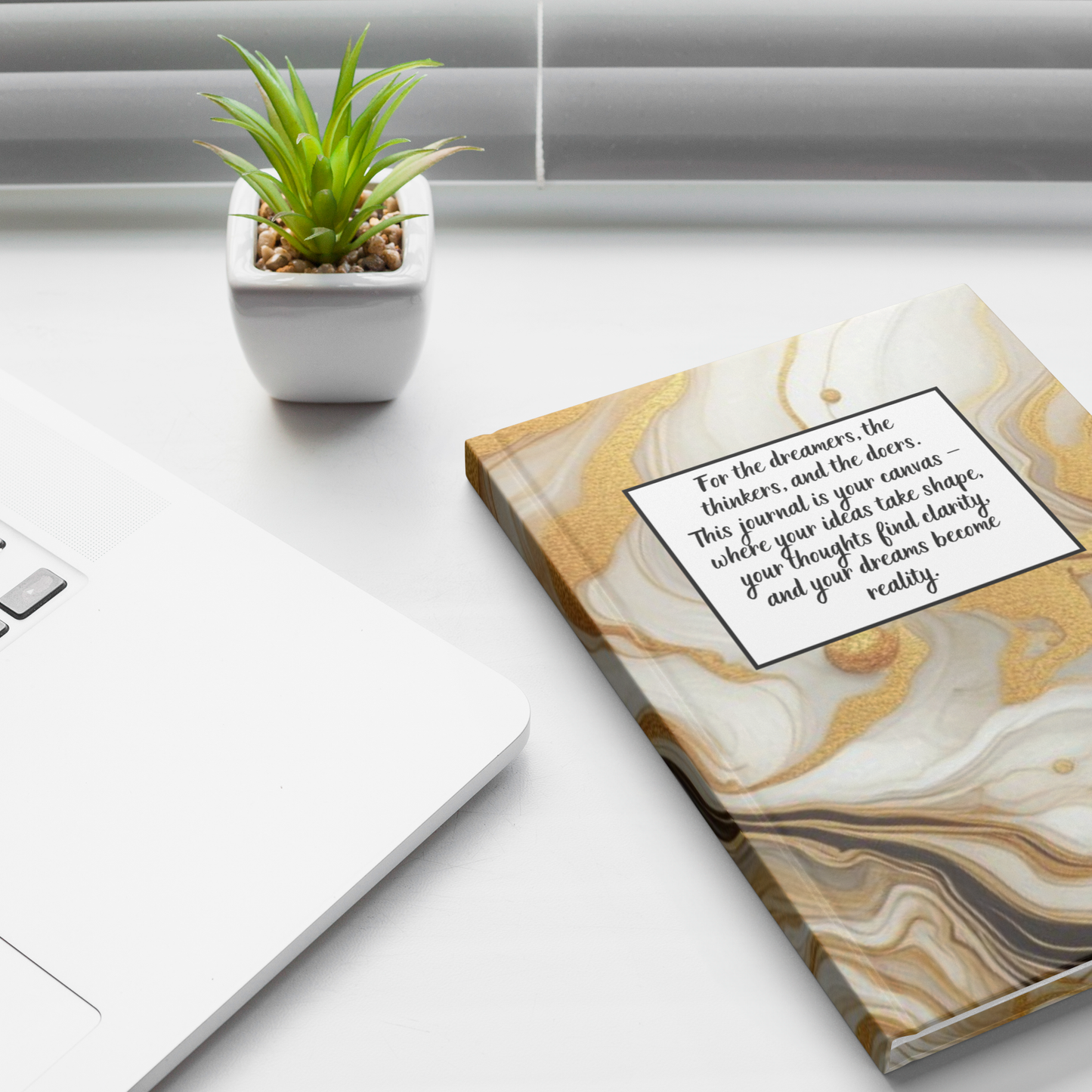 Marble-patterned notebook with inspirational quote on cover next to white laptop and small green plant.