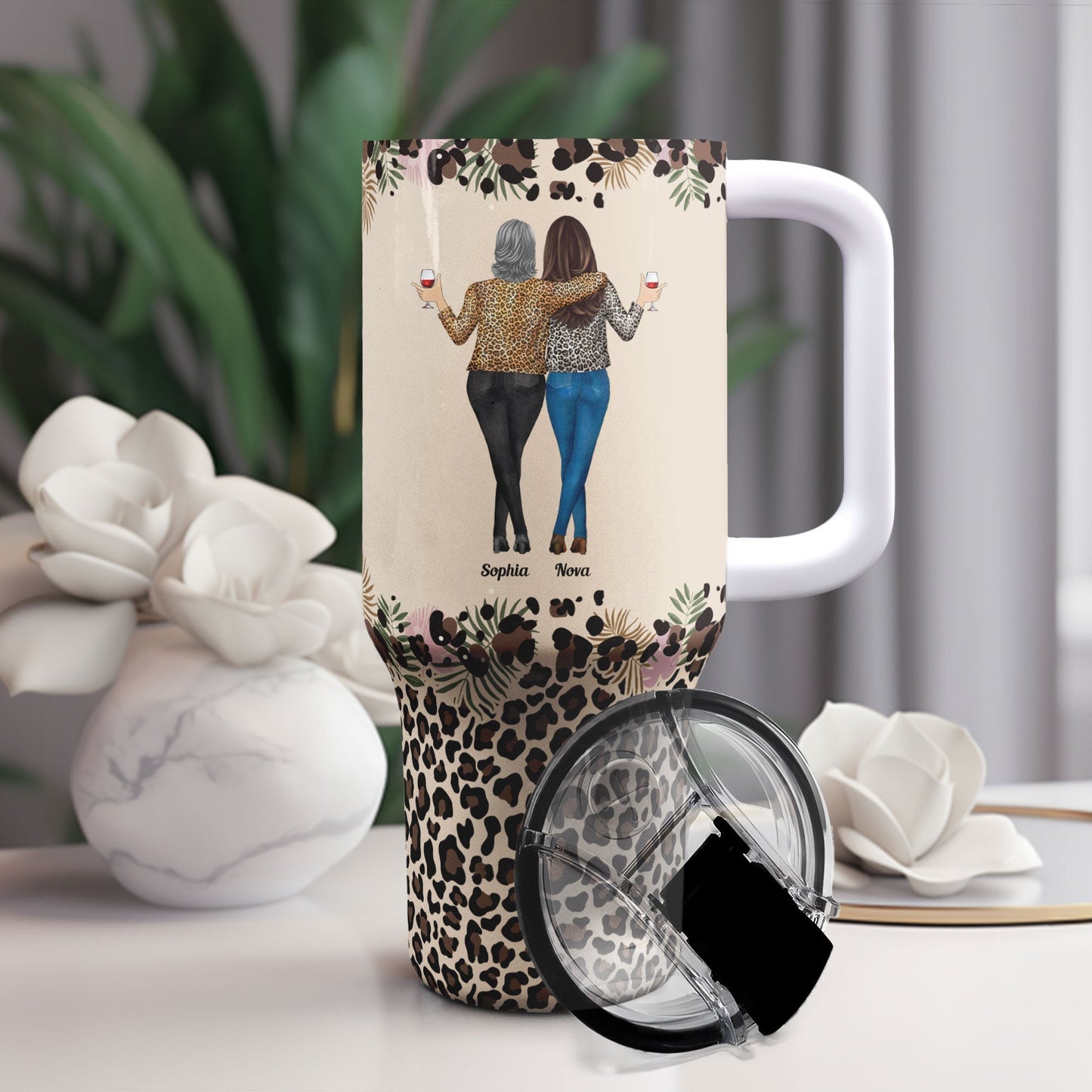 Tumbler with leopard print and personalized cartoon of two women with wine glasses, set on a table with flowers.