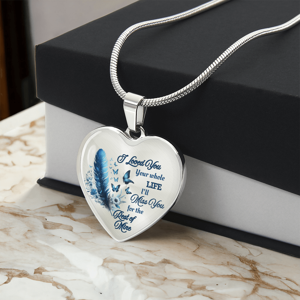Silver heart-shaped pendant necklace with blue feather engraving and cursive letter detail on a marble surface.