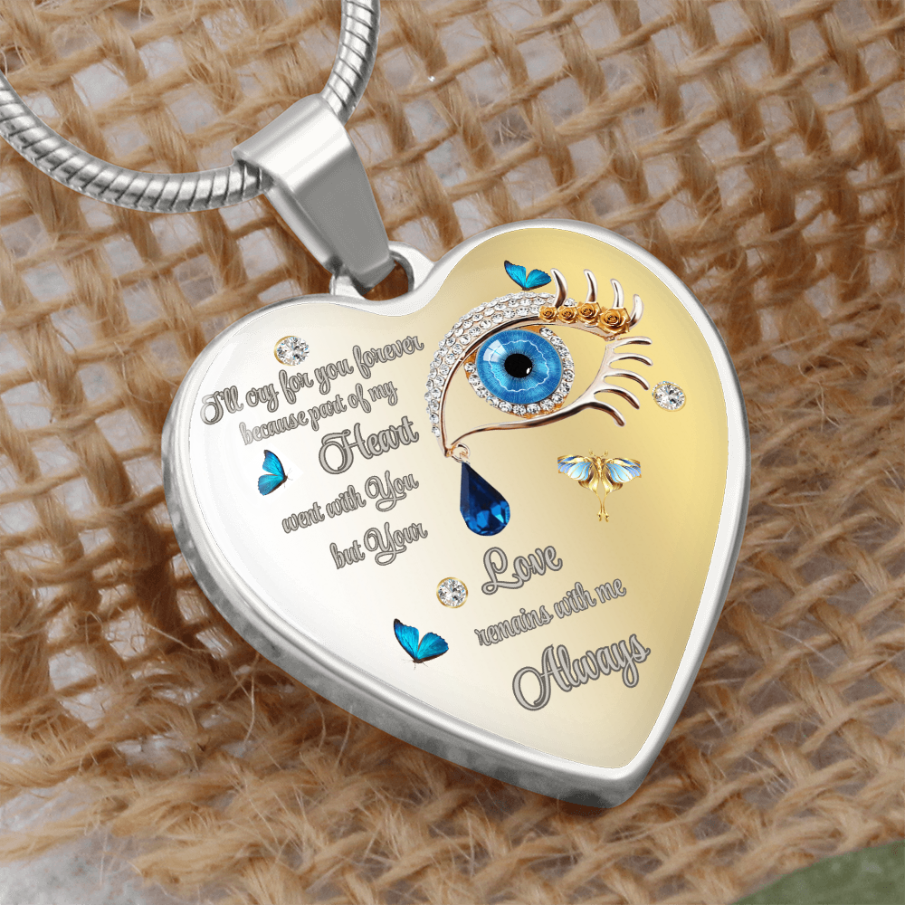 Close-up of a heart-shaped pendant necklace with an ornate blue eye design, gold accents, butterfly motifs, and an engraved inscription on a textured beige backdrop.