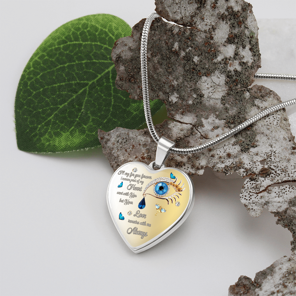 Close-up of a heart-shaped pendant with a silver chain, featuring a blue eye design with butterfly accents and the words "I'll cry for you forever, because part of my heart went with you but your Love remains with me Always," set against a textured stone and a green leaf.
