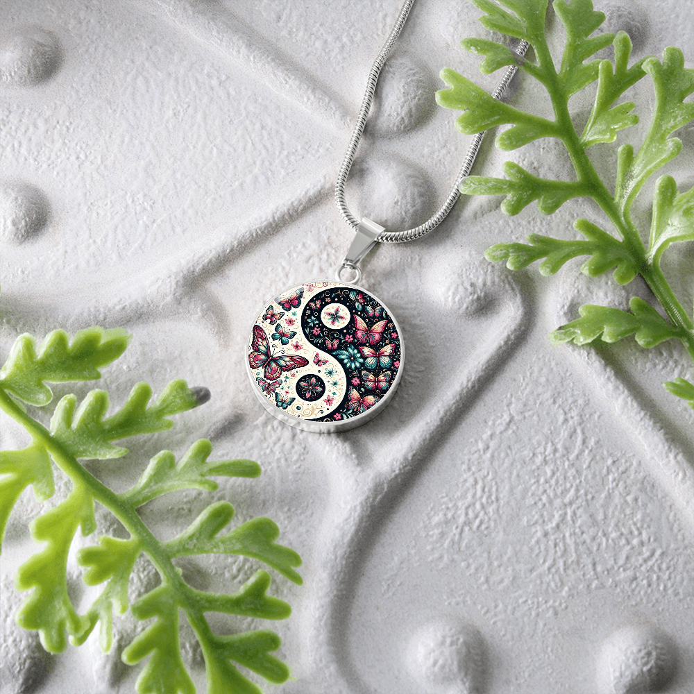 Yin yang butterfly pendant necklace on textured white surface with green plants.
