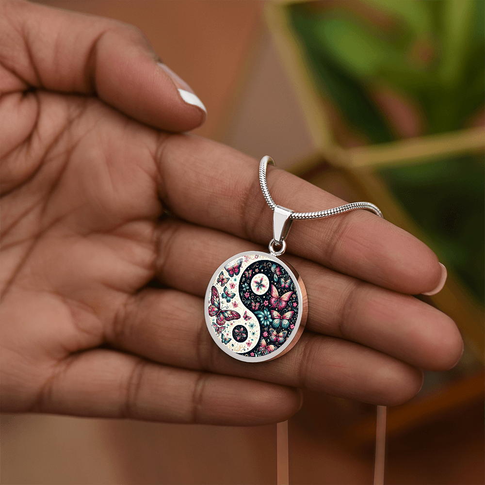 A person holds a silver necklace featuring a circular pendant with a yin-yang symbol filled with butterflies and flowers.