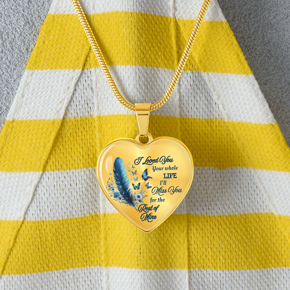 Gold heart-shaped pendant necklace with blue feather and engraved sentimental quote on striped yellow and white fabric.