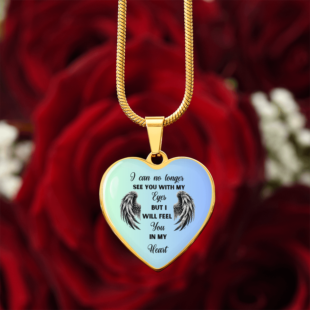 Close-up of a gold heart-shaped pendant necklace with angel wings and the inscription "I can no longer see you with my eyes, but I will feel you in my heart" against a blurred background of red roses.
