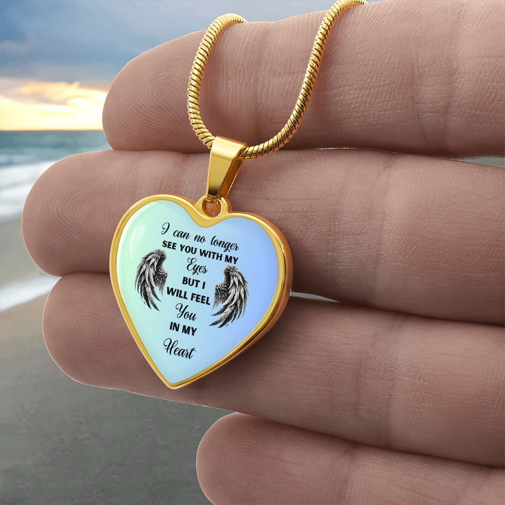 Heart-shaped pendant necklace with a gold chain and bezel, displaying the text "I can no longer see you with my eyes, but I will feel you in my heart" with angel wings on a blue and white background, held by a hand against a beach backdrop.