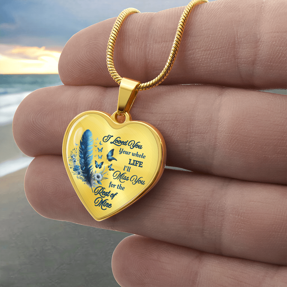 Gold heart-shaped pendant necklace with blue feather and engraved sentimental message held between fingers.