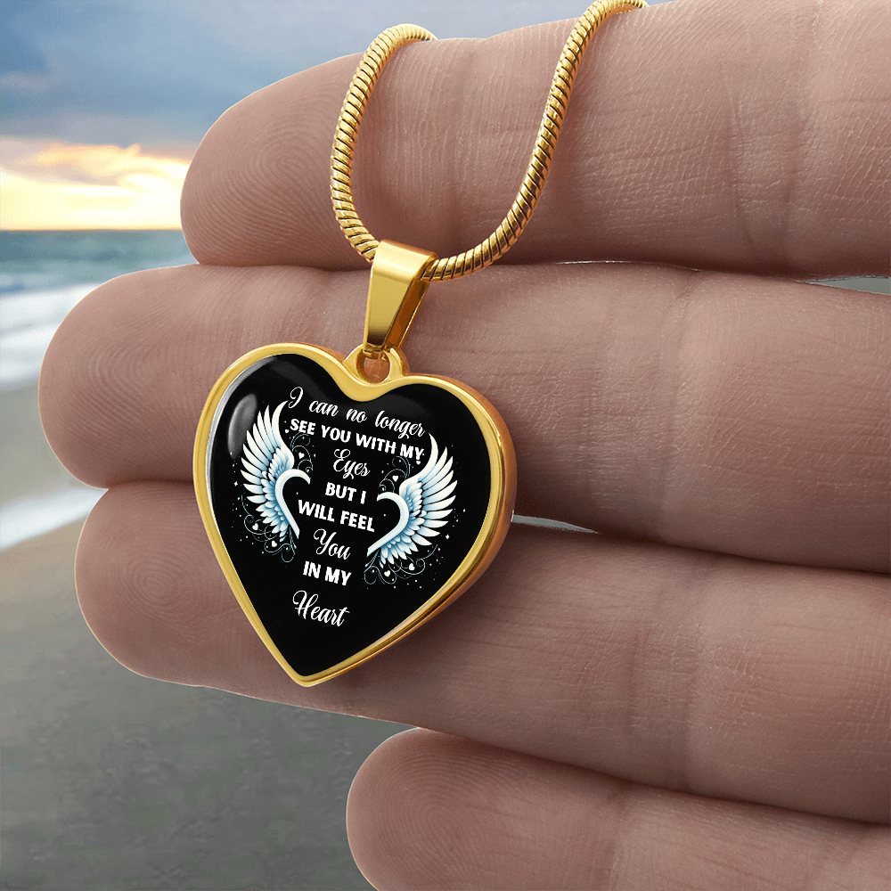 Close-up of a gold heart-shaped pendant necklace held against a beach background. The pendant has the words, "I can no longer SEE YOU WITH MY Eyes BUT I WILL FEEL You IN MY Heart" and a pair of white angel wings printed on it.