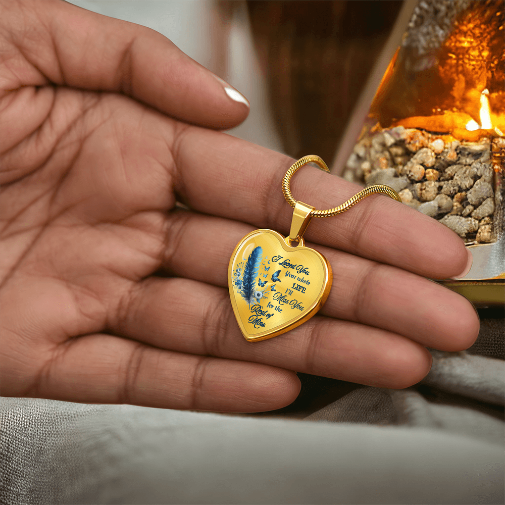 Gold heart-shaped pendant necklace with blue feather and engraved letter detail held in hand.