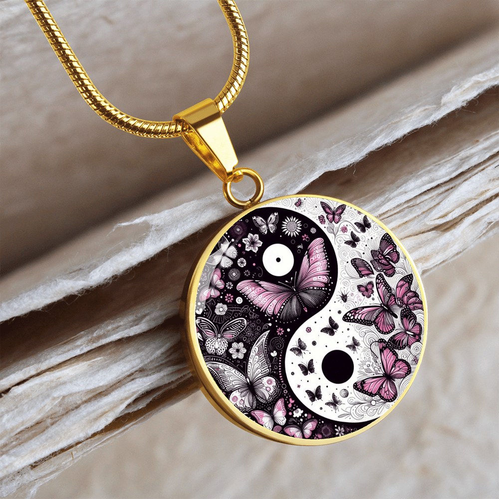A gold-colored necklace with a round yin-yang pendant. The pendant features a butterfly design in pink, white, and black. The pendant is resting on a light-colored, textured surface.
