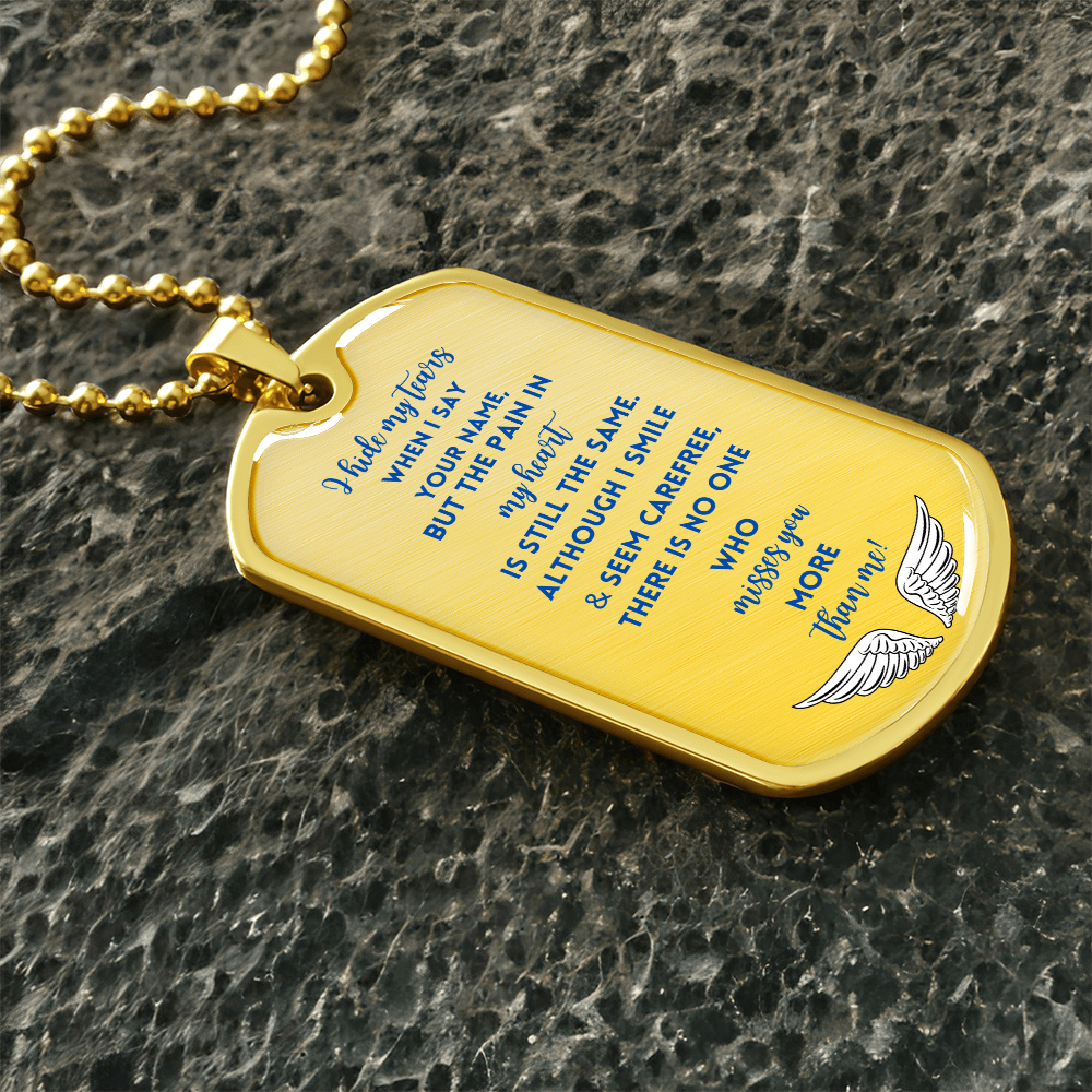 Gold memorial dog tag necklace with engraved blue text and angel wings on textured dark surface.
