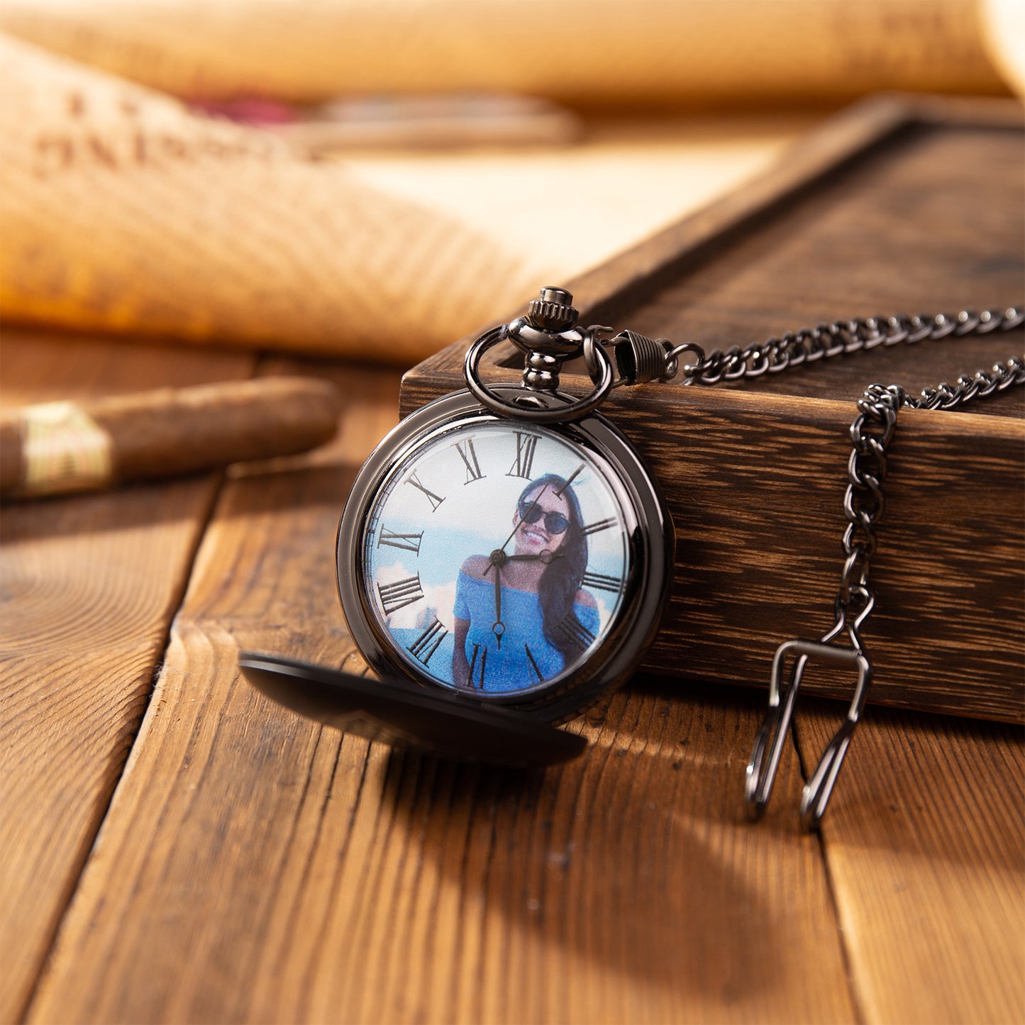 Black pocket watch with Roman numerals and custom photo on the dial, attached to a chain on wooden surface.