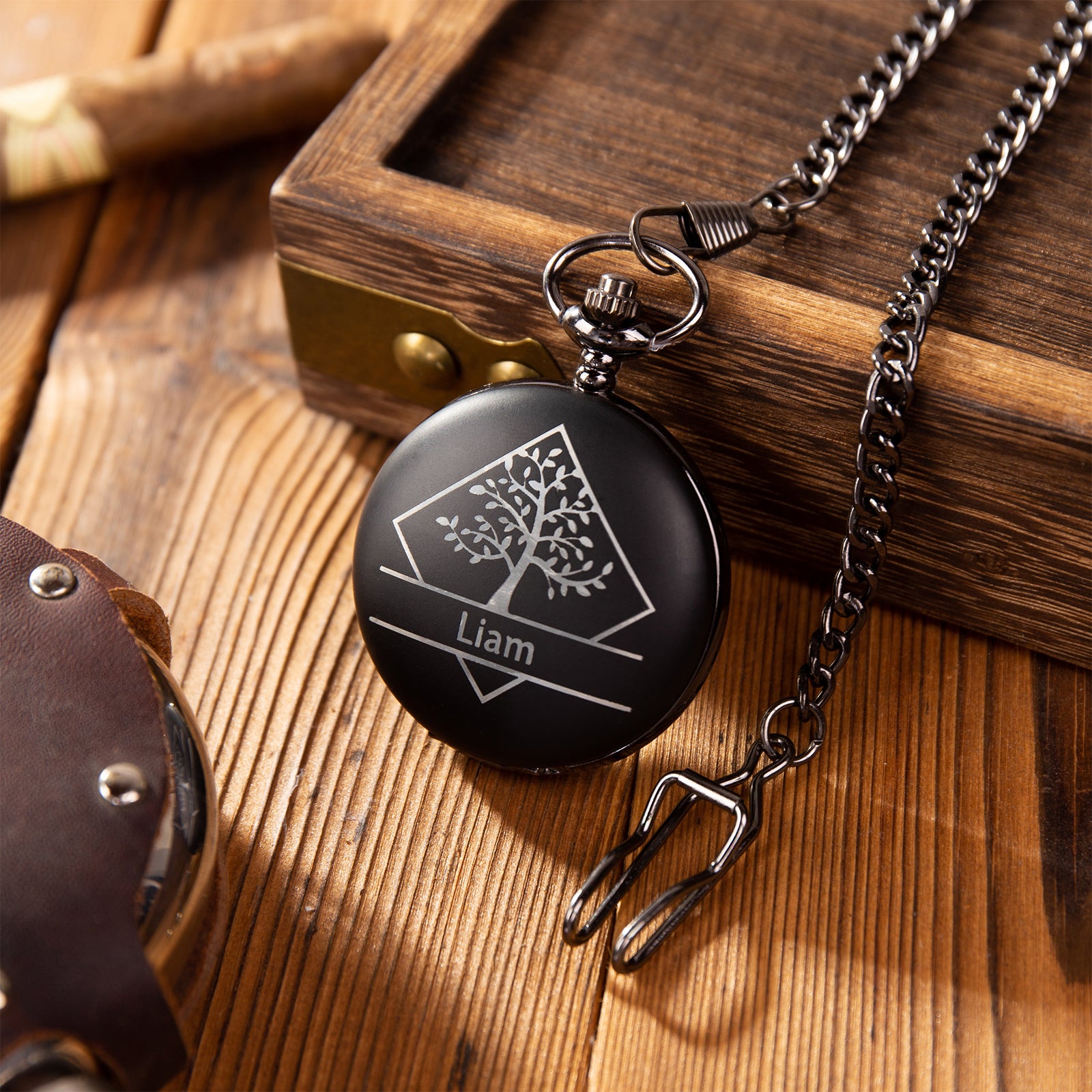 Black pocket watch with engraved tree design and name "Liam" on wooden surface with chain.