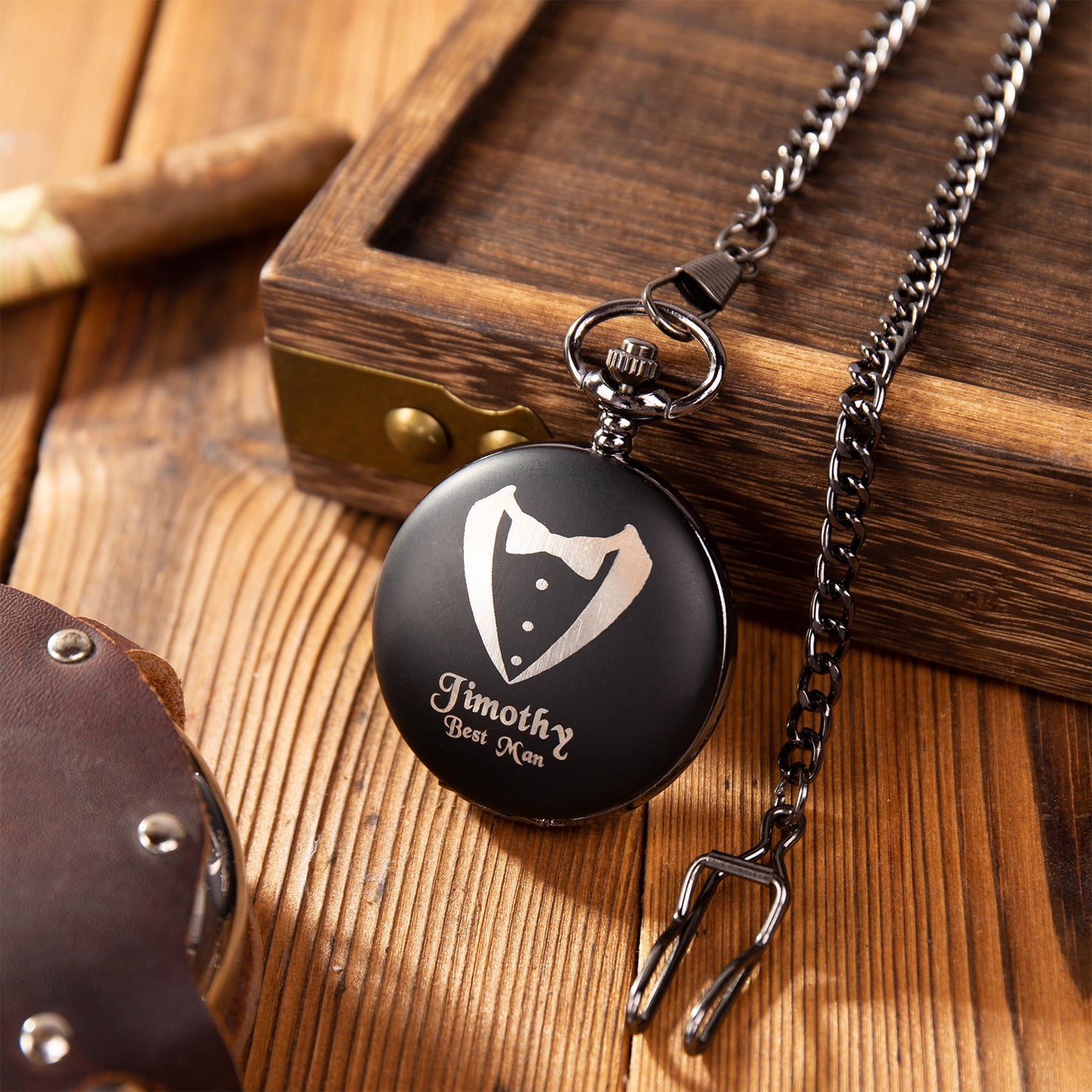 Black metal pocket watch with tuxedo design and personalized name on wooden surface with chain.