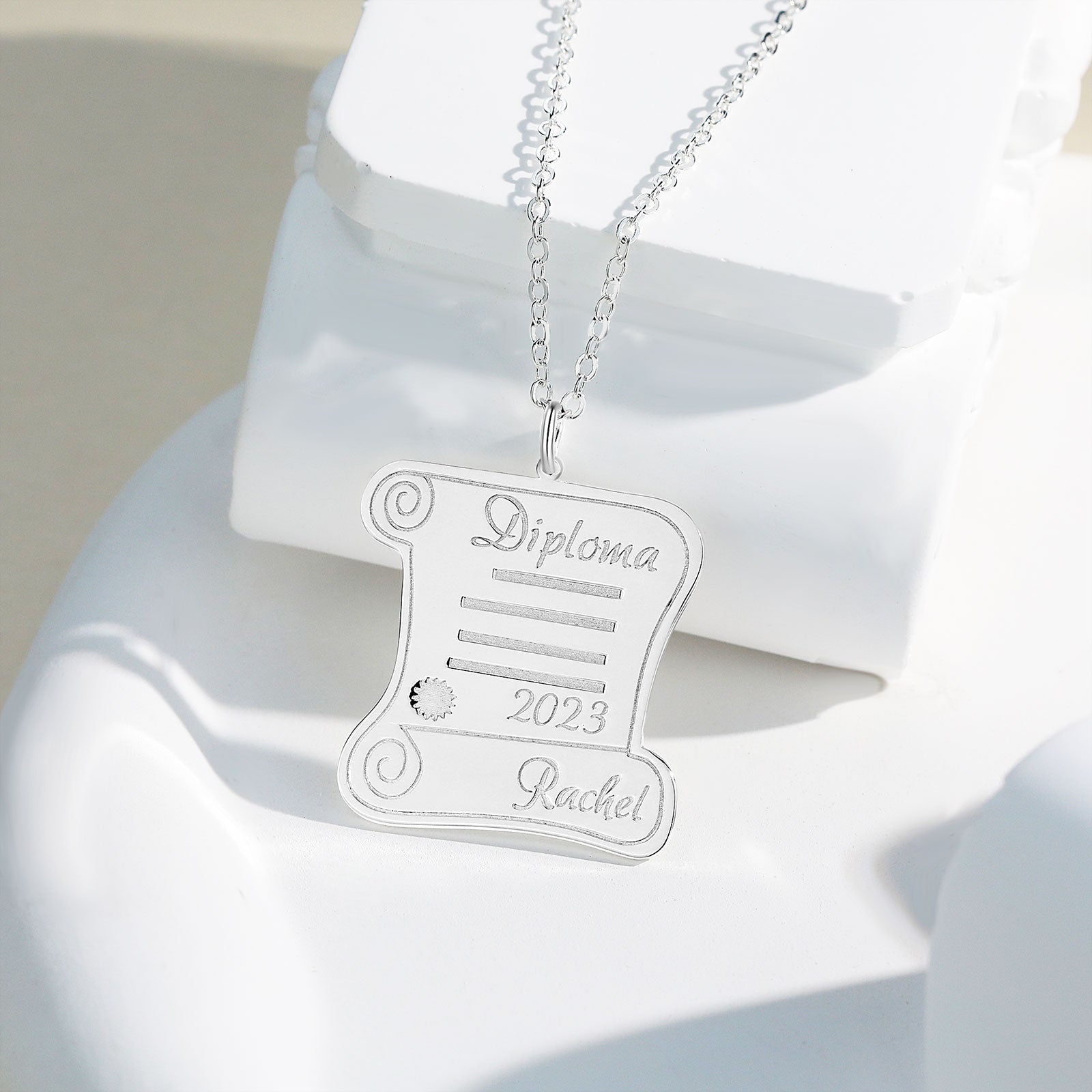 Silver pendant necklace shaped like a scroll with engraved "Diploma," "2023," and personalized name on a silver chain.