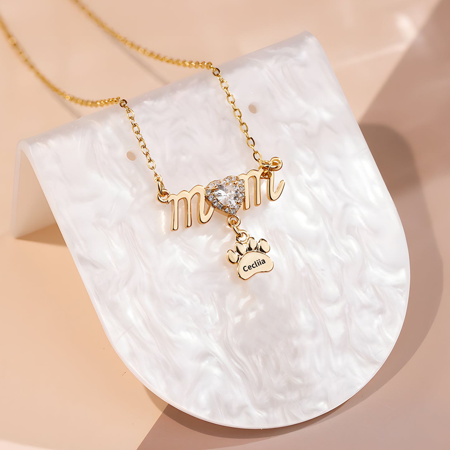Gold necklace with "mom" script, crystal heart, and small paw charm engraved "Celia" on a pearly white stand.