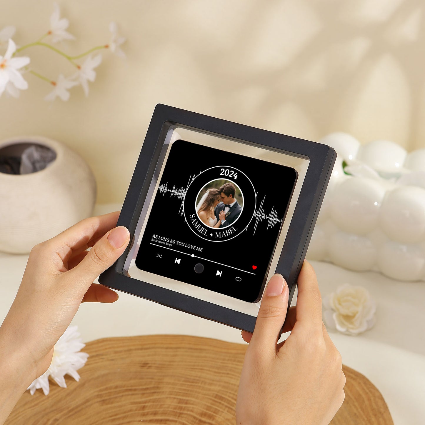 A woman holds a personalized music plaque in a black frame, featuring a wedding photo within a waveform design, along with text indicating names, a year, and a song title.