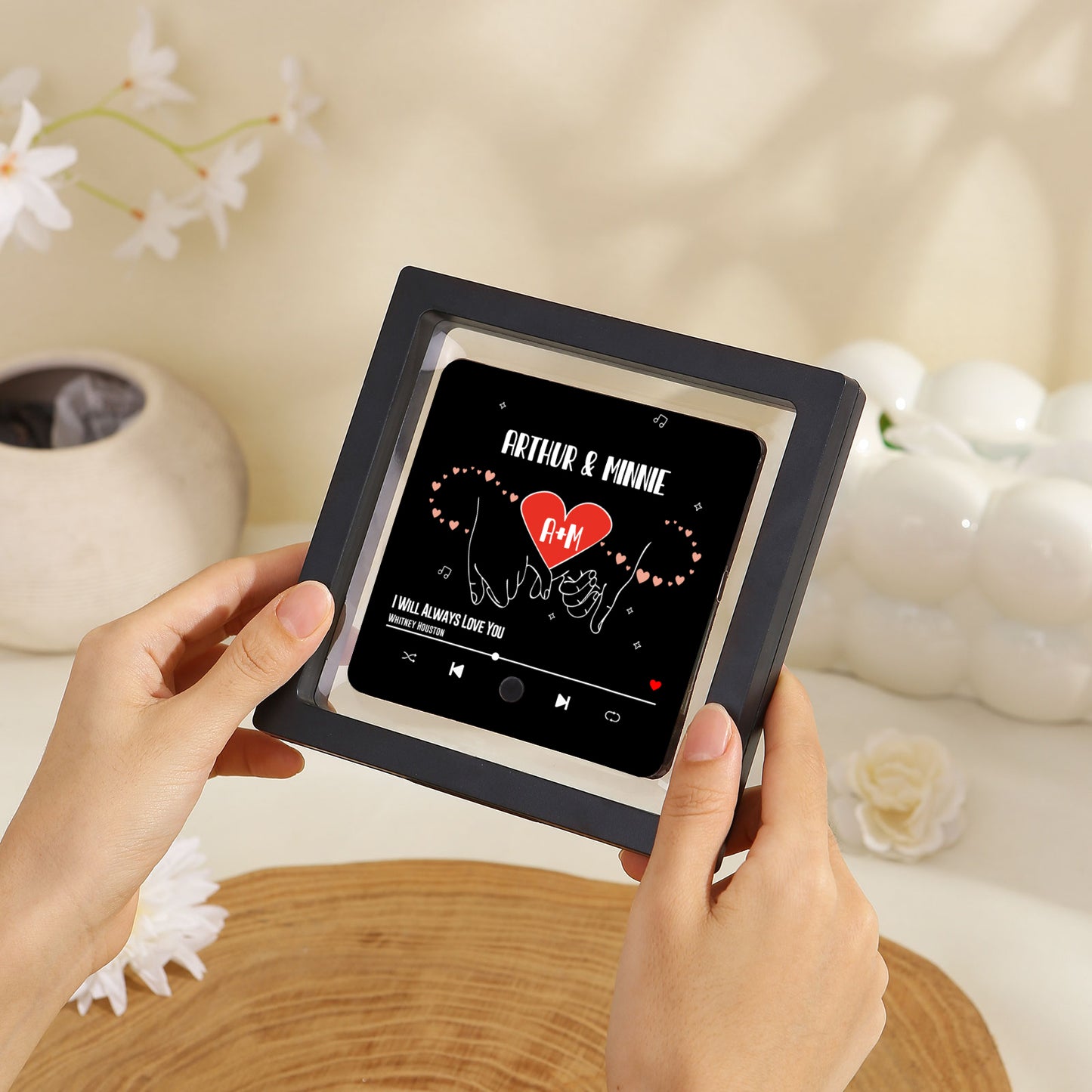 A framed music art display is held by two hands, featuring a black background with the names "Arthur & Minnie" above a red heart displaying "A+M" on it.