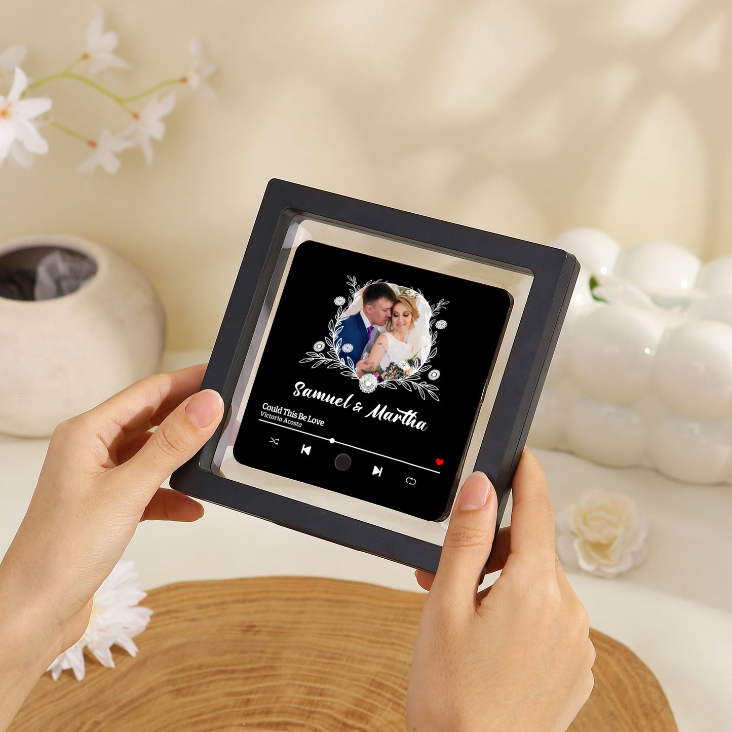Close-up of hands holding a framed personalized photo art with a black frame, black background, white decorations, and an image of a couple inside, with song title "Could This Be Love".