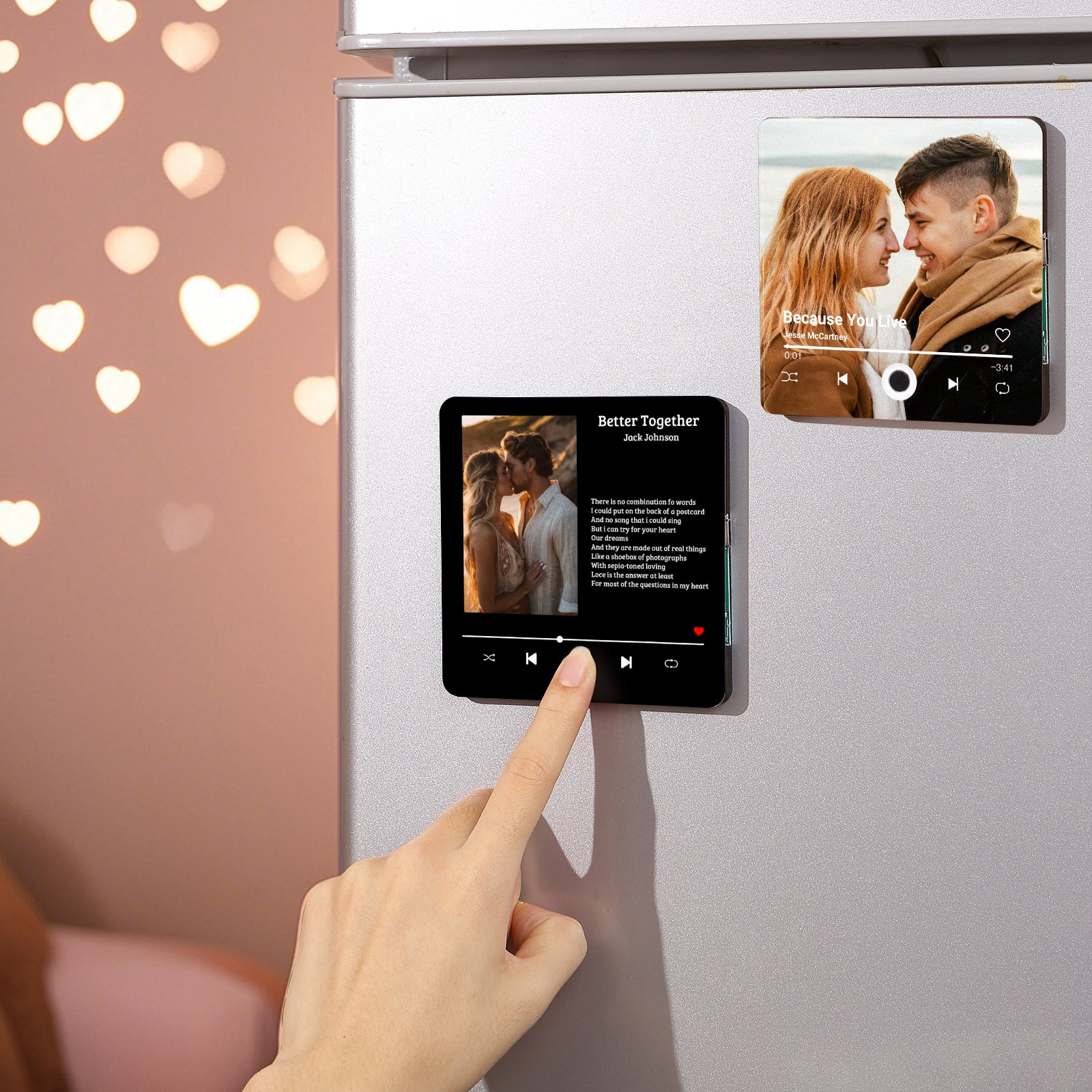 Close up of two magnetic, personalized square tiles on a silver refrigerator. On the left, a hand touches one magnetic tile depicting a couple kissing with the song "Better Together" by Jack Johnson. On the right, another magnet tile shows a couple looking at each other with the song "Because You Live" by Jesse McCartney. Soft, out of focus, heart-shaped lights on the left.