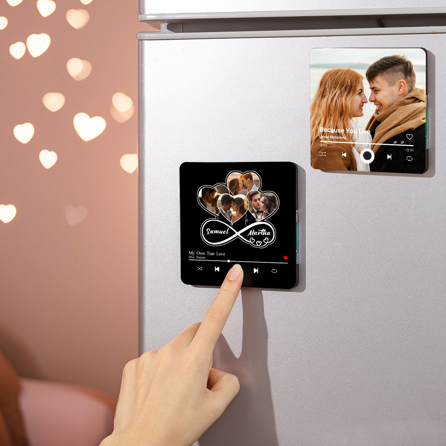 Close-up of a silver refrigerator with two photo-decorated magnets. A hand touches the bottom magnet, which has five heart-shaped couple photos, an infinity symbol, the names "Samuel" and "Martha", and music player icons. The top magnet features a couple facing each other and the song "Because You Live". The background includes a pink wall with heart-shaped bokeh.
