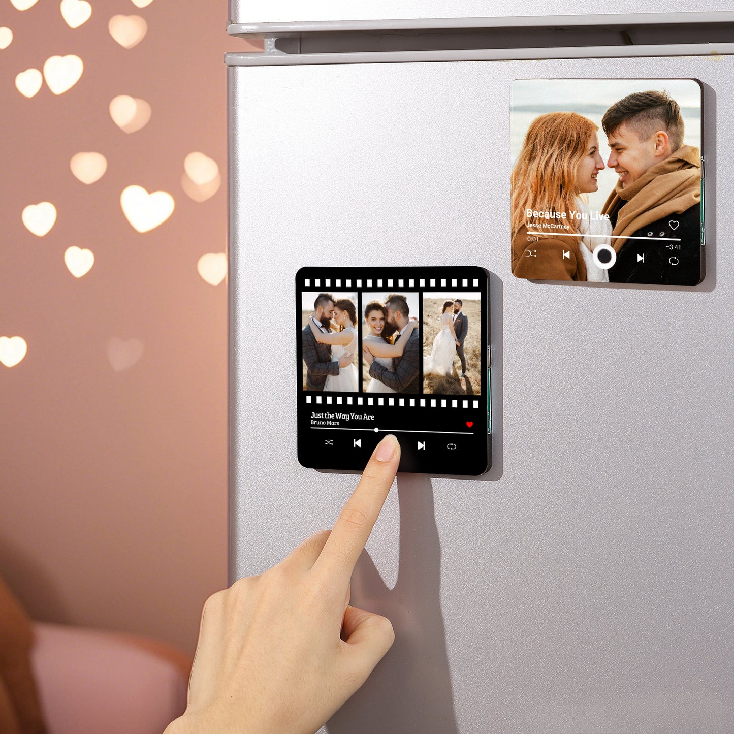 Finger pressing on a black and white photo collage magnet resembling a music player on a silver refrigerator, next to another photo magnet, with blurred heart-shaped lights in the background.