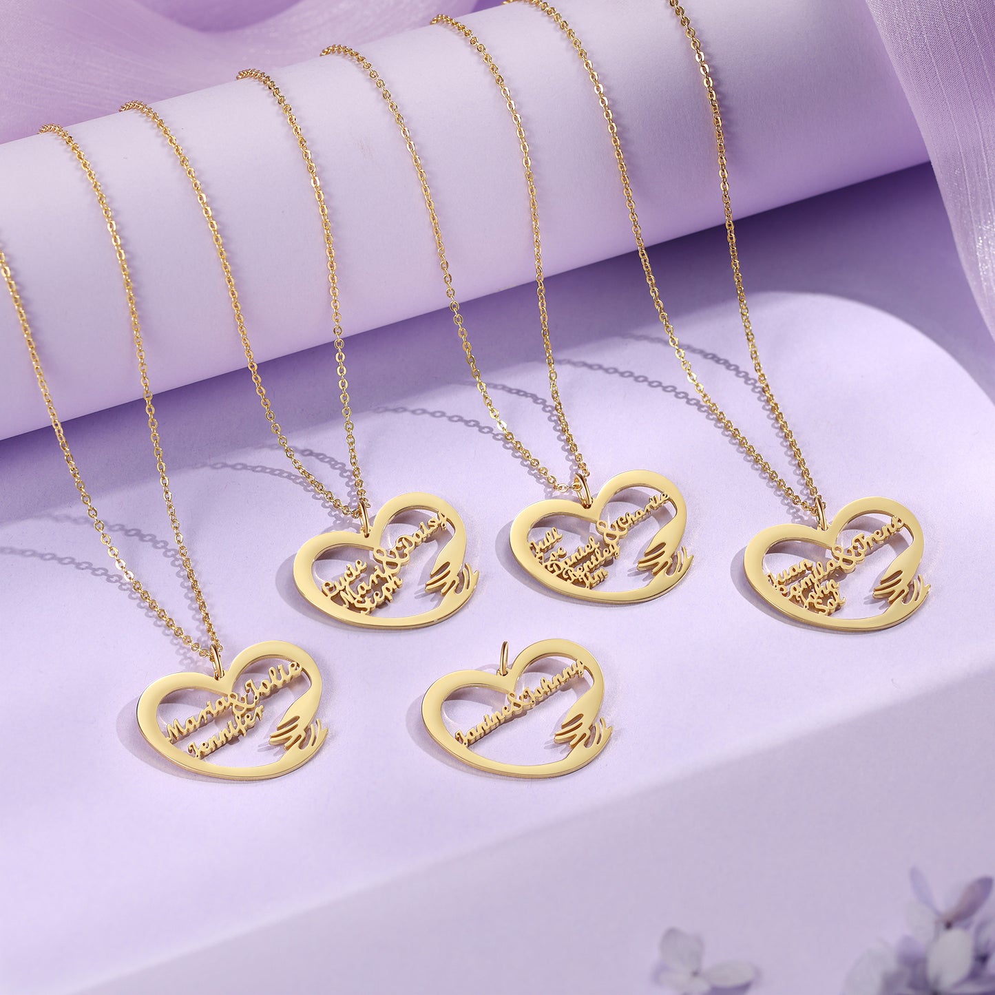 Gold plated heart-shaped necklaces with custom name engraving on delicate chains on lavender fabric background.
