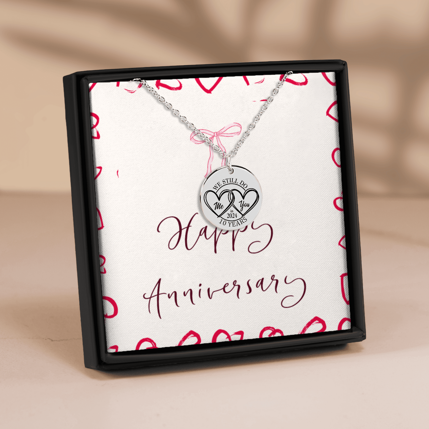 A silver anniversary necklace with a round pendant engraved with '10 YEARS' and two interlocking hearts, presented in a black gift box with a white insert that reads 'Happy Anniversary' and decorated with red hearts.