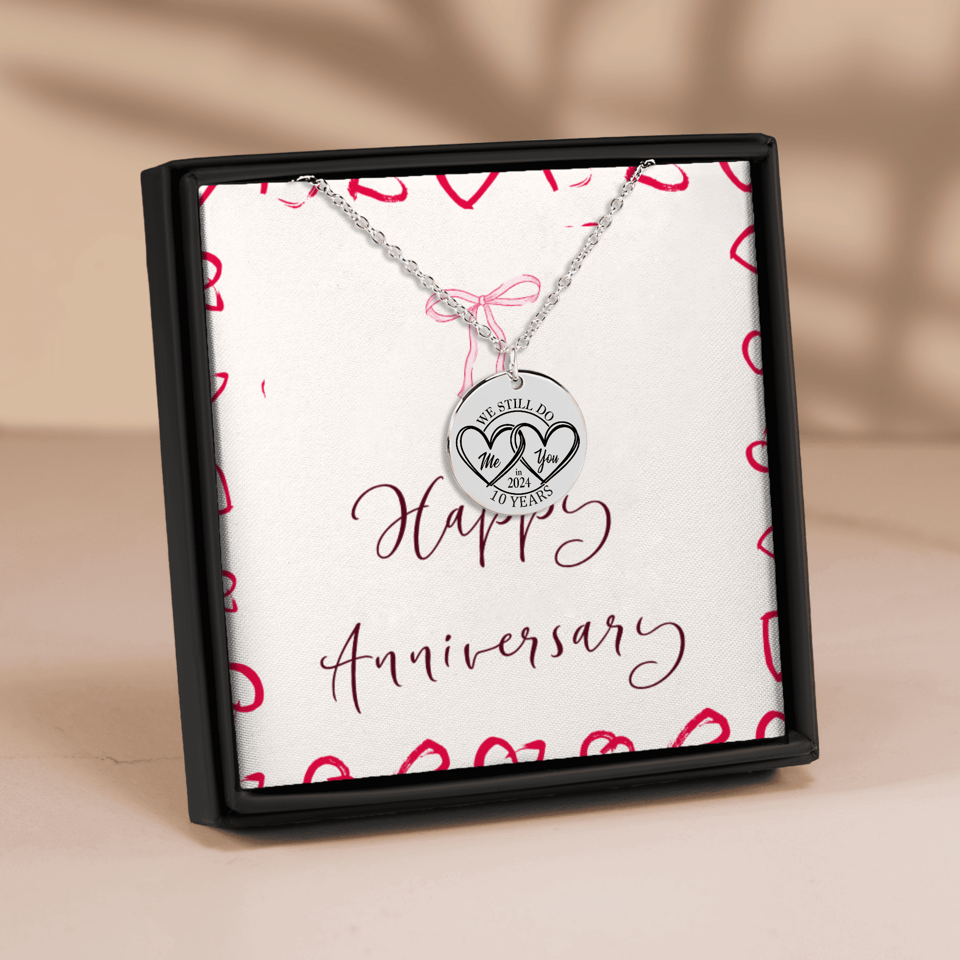 A silver anniversary necklace with a round pendant engraved with '10 YEARS' and two interlocking hearts, presented in a black gift box with a white insert that reads 'Happy Anniversary' and decorated with red hearts.