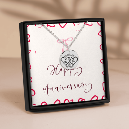 A silver anniversary necklace with a round pendant engraved with '10 YEARS' and two interlocking hearts, presented in a black gift box with a white insert that reads 'Happy Anniversary' and decorated with red hearts.